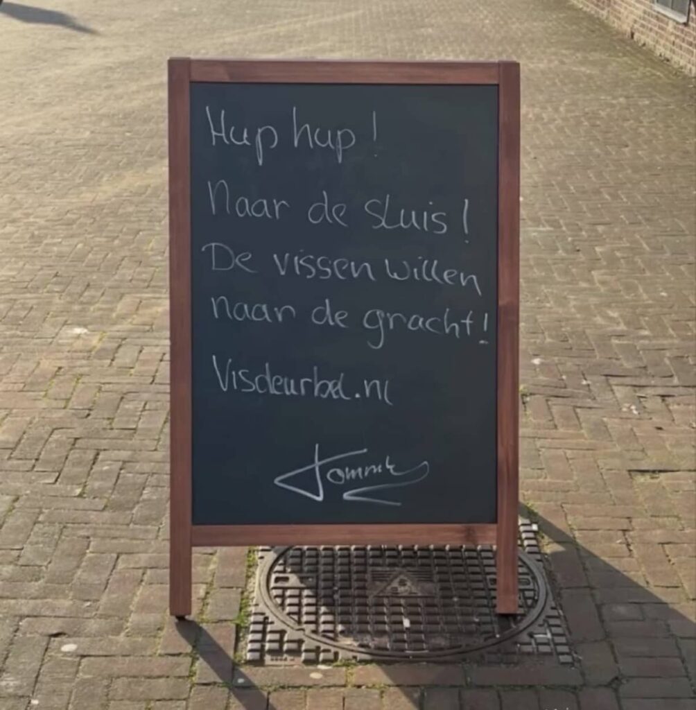Chalkboard on a street with the text: “Come on, to the lock! The fish want to reach the canal! visdeurbel.nl – Tommy.”